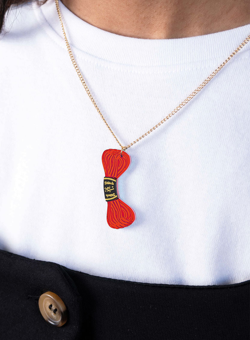A close-up of a person wearing a gold chain necklace with a Tatty Devine red embroidery thread pendant, styled over a white t-shirt and black top.