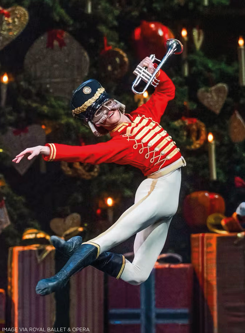 A dancer in a Nutcracker-style holiday nutcruppe costume leaps through the air while holding a silver trumpet. The costume features a bright red jacket with gold stripes and trim, white pants, dark blue boots, and a dark blue hat with gold detailing.
