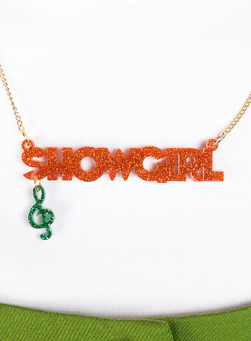 Tatty Devine Name Necklace with 'SHOWGIRL' in orange glitter and green pendant on a white background.